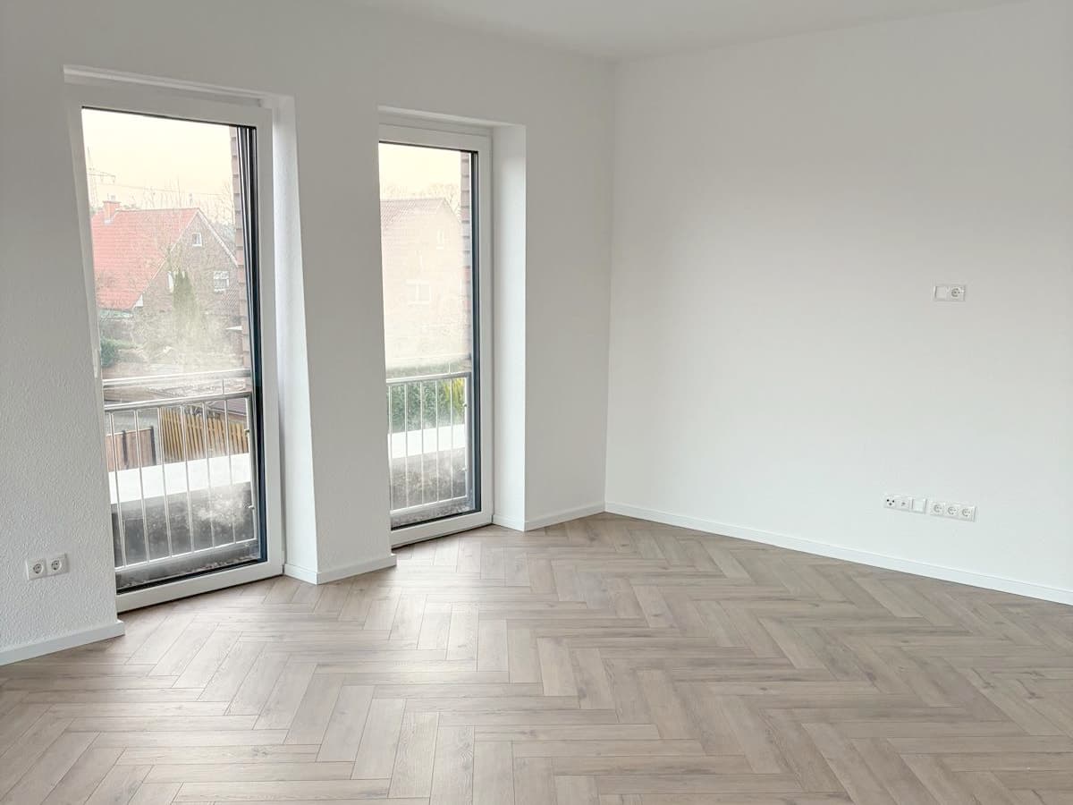 Leerer, frisch sanierter Wohnraum mit hellem Fischgrätboden und zwei bodentiefen Fenstern zum Balkon
