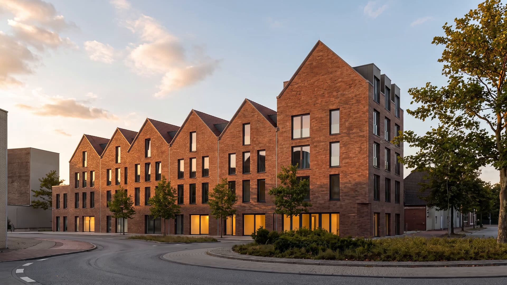 Backsteingebäude mit einer Reihe spitzer Giebel im Abendlicht, warm erleuchtete Fenster im Erdgeschoss, davor eine leere Straße
