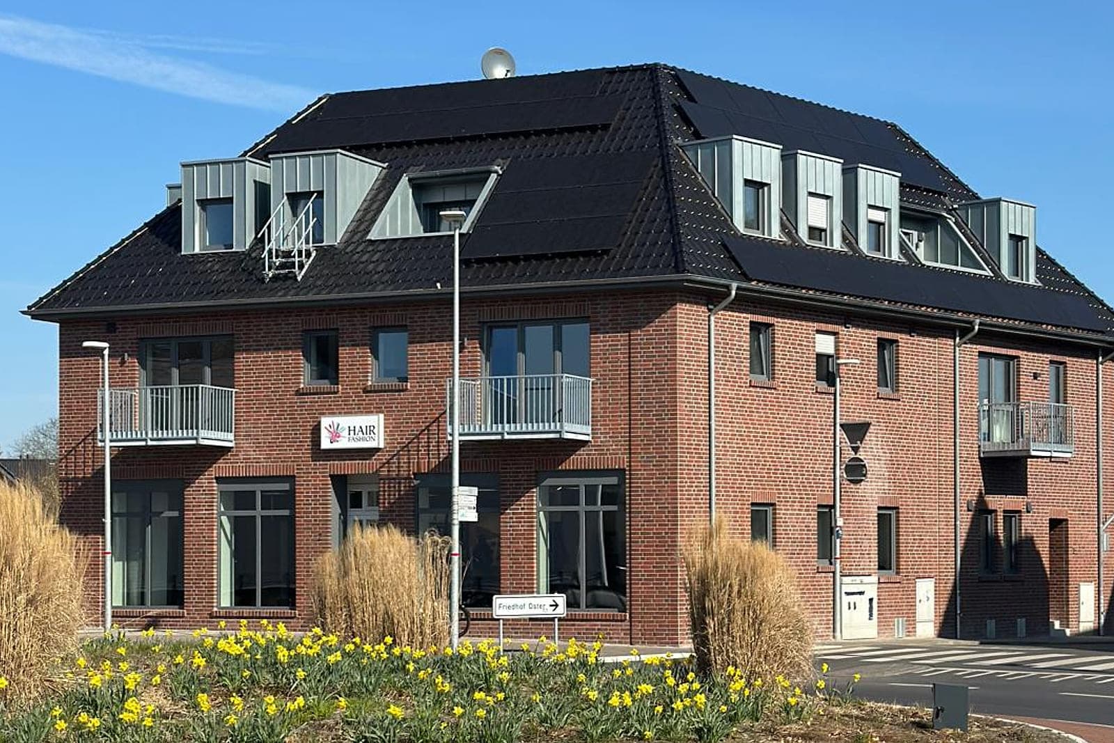 Dasselbe Gebäude nach dem Umbau: rote Klinkerfassade, neu gedecktes dunkles Dach mit breiten Gaubenbändern, Balkone mit Metallgeländer, davor der blühende Kreisverkehr