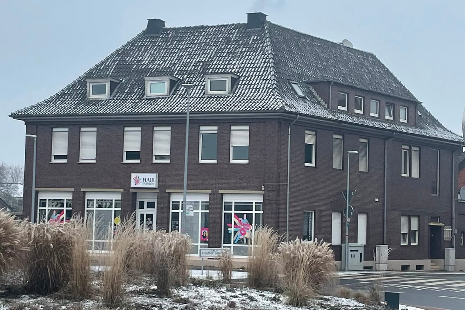 Das Gebäude in Ochtrup vor dem Umbau: dunkel verputzte Fassade mit hellem Erdgeschoss, altes Ziegeldach mit drei kleinen Gauben, davor der Kreisverkehr im Winter