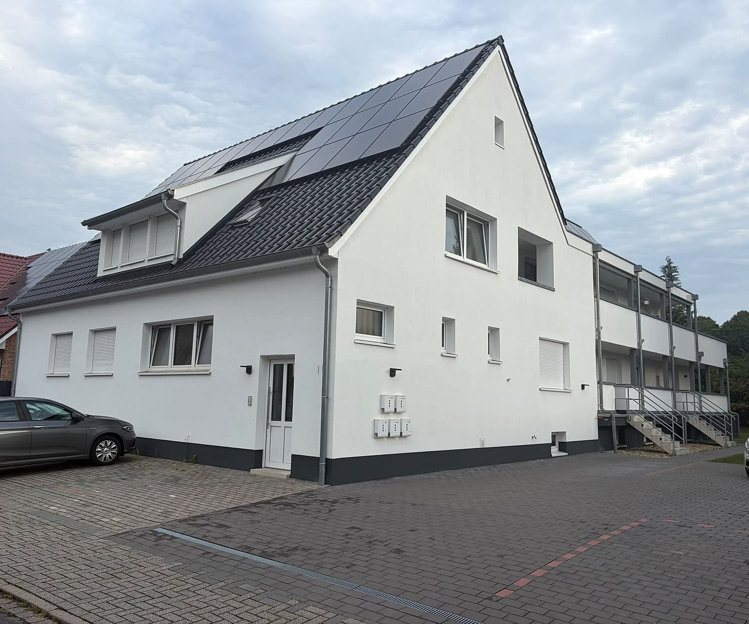 Dasselbe Gebäude nach der Sanierung: weiß verputzt, dunkles Dach mit Photovoltaik und Gauben, rechts ein außenliegender Laubengang über zwei Geschosse